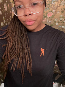 A black woman with clear framed glasses, sporting with a dark grey long sleeve shirt with an orange embroidered silhouette of "stronger" in ASL, poses for a selfie 